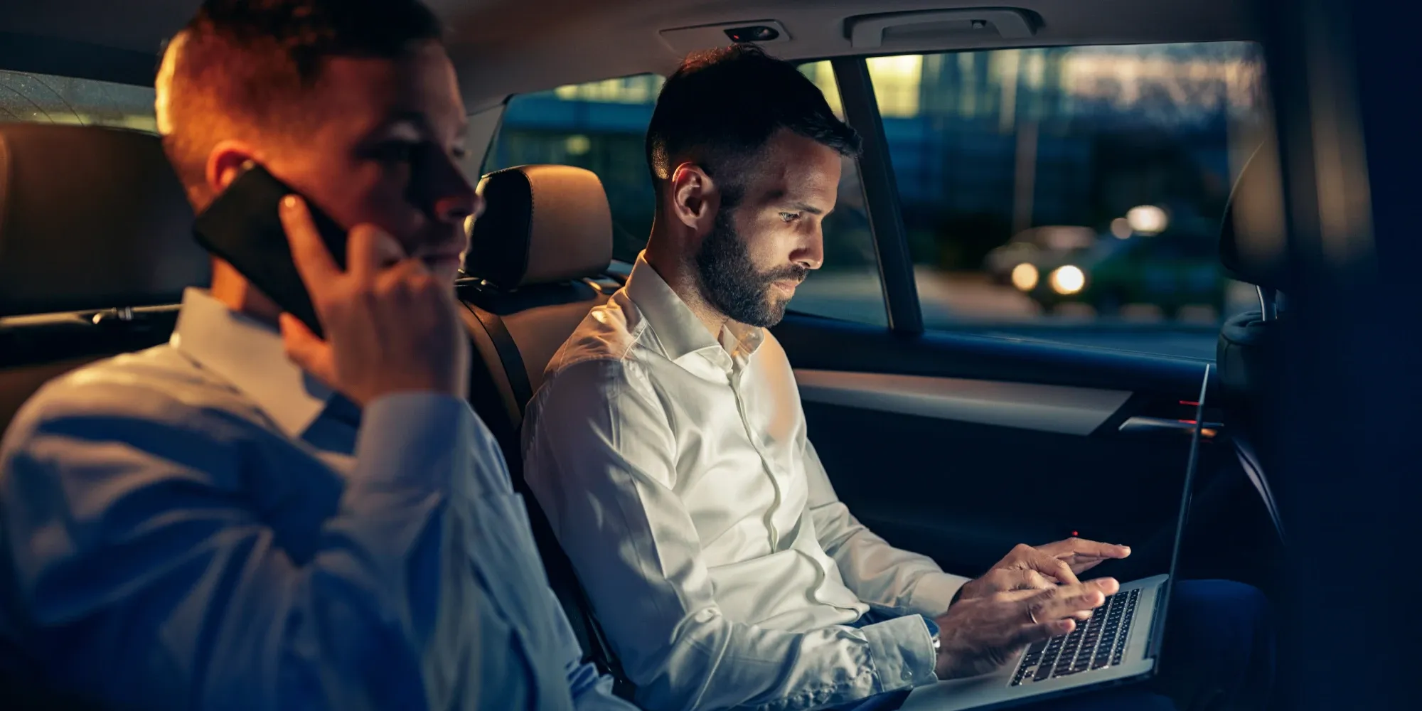 Cover image for "The Human Engine Behind Autonomous Systems." Two businessmen work productively in the backseat of a moving car at night; one talks on a smartphone in the foreground while the other types on a laptop.