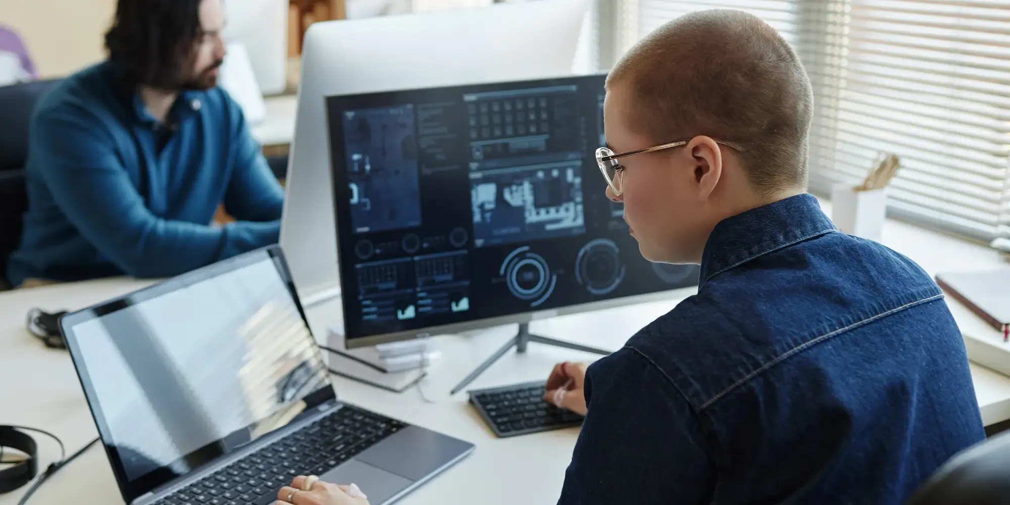 An analyst with a buzzed haircut and glasses uses multiple screens to analyze complex data in a bright, modern office.