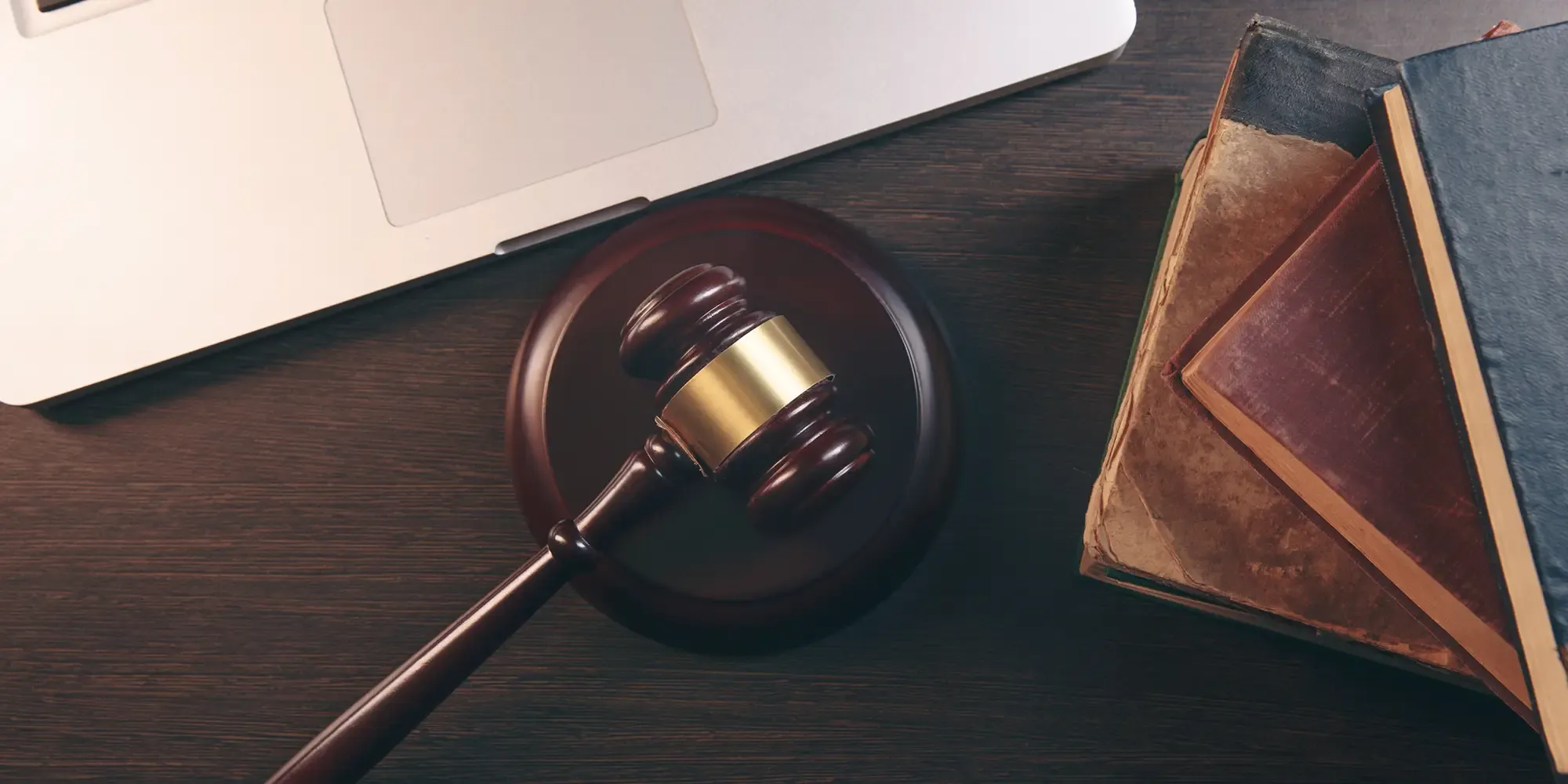 "A top-down view of a wooden gavel, a modern silver laptop, and a stack of old reference books resting on a dark wooden desk, symbolizing the intersection of legal regulations, traditional policies, and modern business strategy involved in Governance, Risk, and Compliance (GRC)