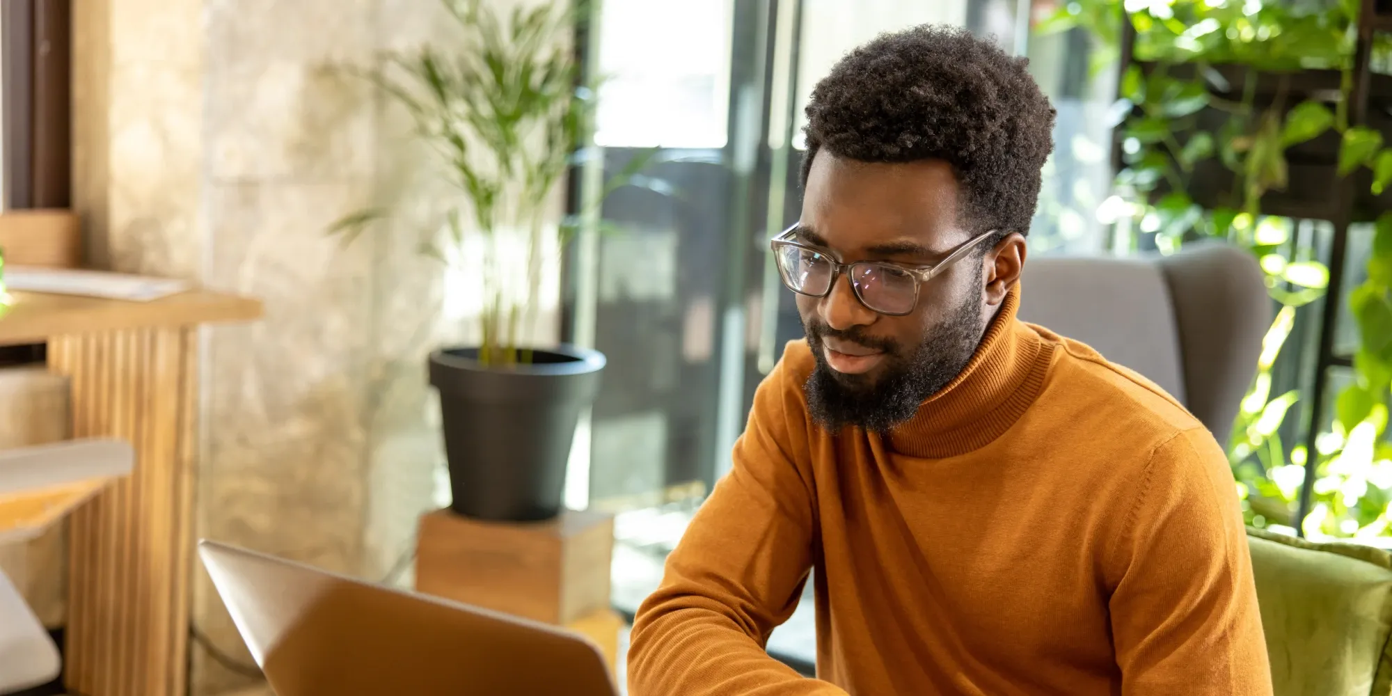 A focused man with a beard and glasses wearing an orange turtleneck looks at a laptop while working from a modern, sunlit cafe with green plants.