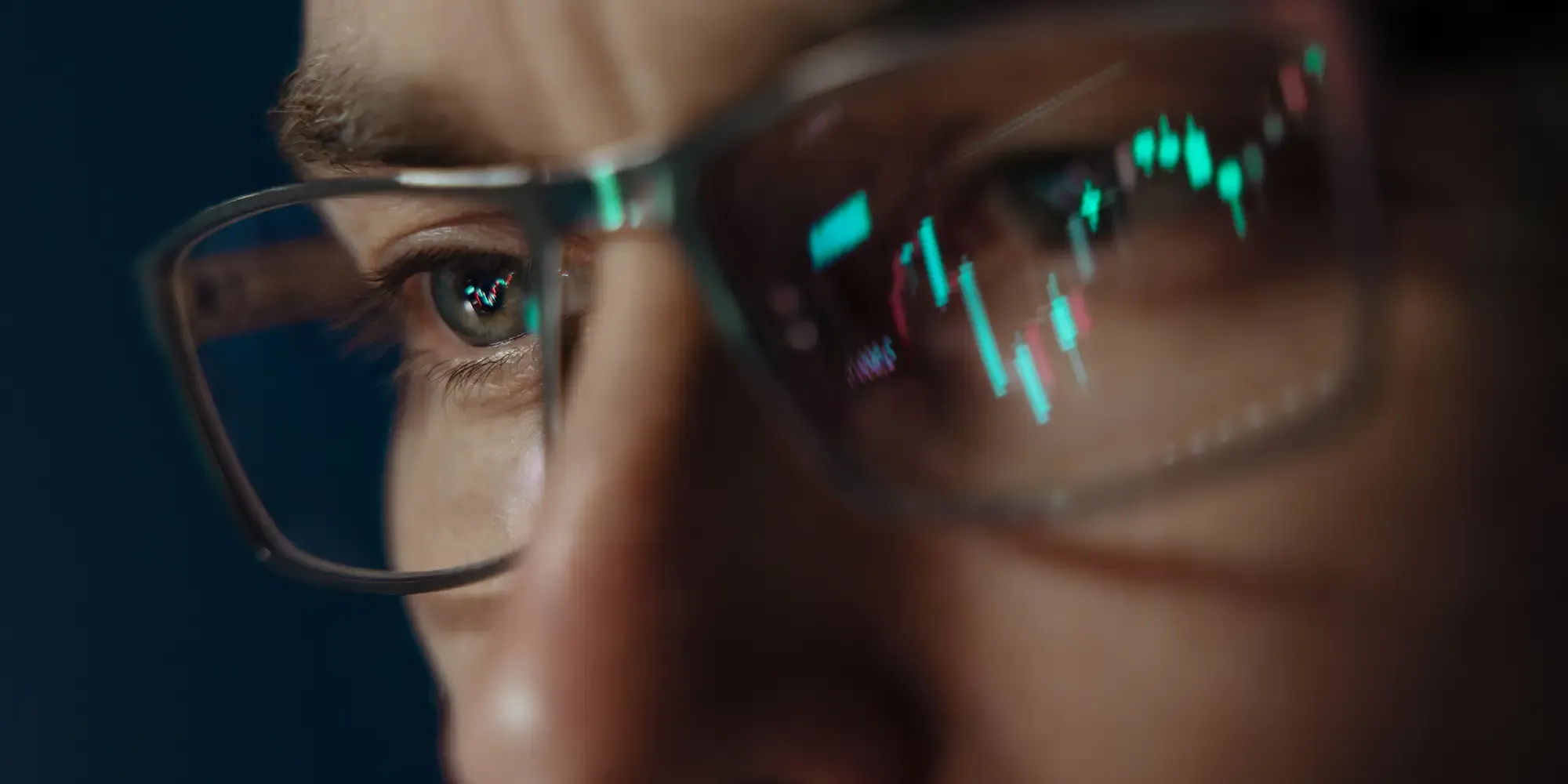 A close-up of a person wearing glasses, with glowing neon green and pink financial candlestick charts reflecting vividly across the lenses and in their eye, illustrating an intense focus on market data.